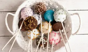 Kolorowe cake pops w białym koszyku, udekorowane posypkami i kuleczkami.