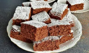 Pyszne ciasto czekoladowe pokrojone w kostkę, posypane cukrem pudrem, ułożone na białym talerzu.
