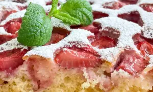 Puszyste ciasto jogurtowe z kawałkami truskawek, posypane cukrem pudrem i ozdobione listkiem mięty.