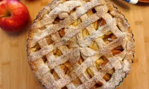 Pyszna tarta z jabłkami, posypana cukrem pudrem, obok leżą świeże jabłka.