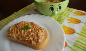Pyszna pasta z makreli i twarogu na kromce chleba, ozdobiona listkiem bazylii. Obok miseczka z pastą i wesoła buźka.