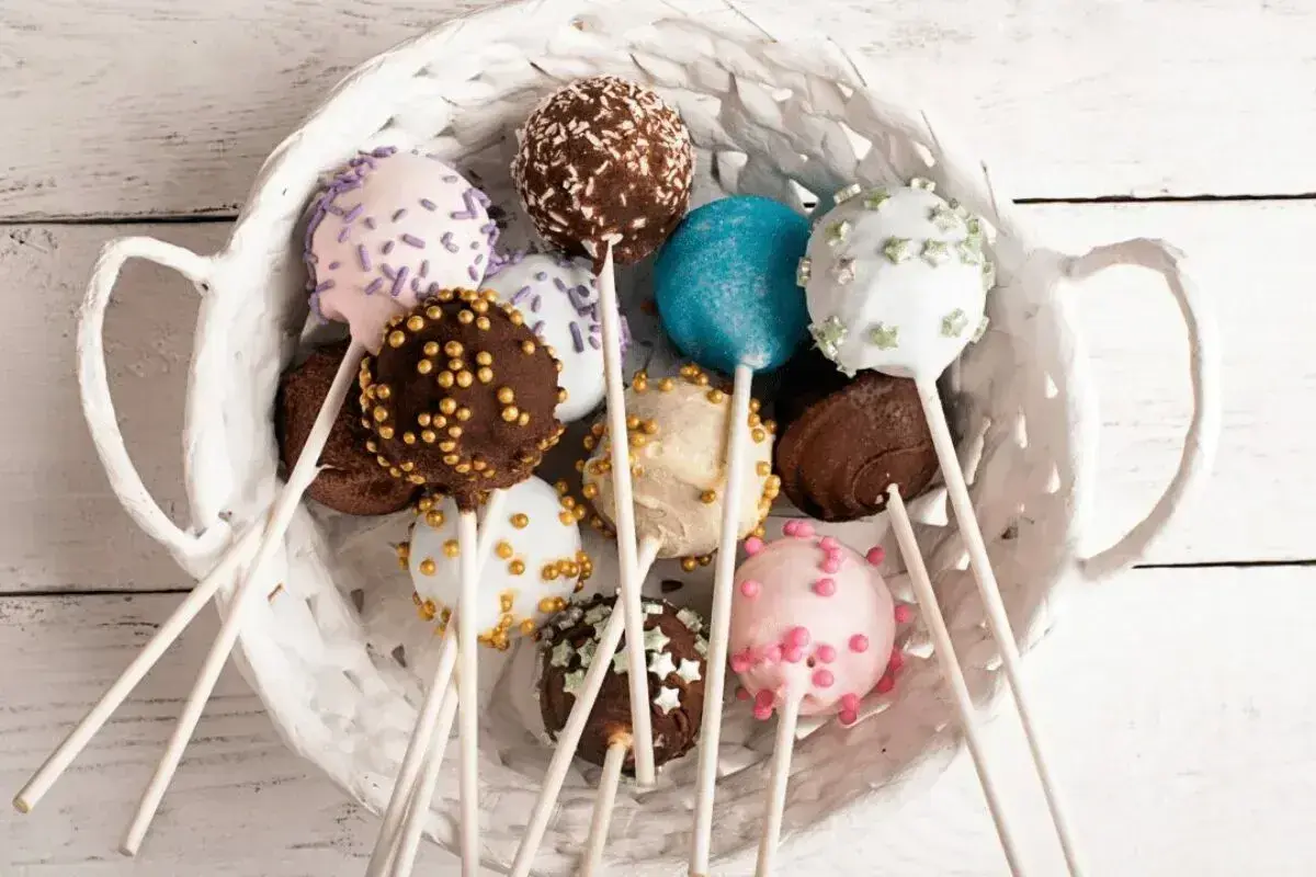 Kolorowe cake pops w białym koszyku, udekorowane posypkami i kuleczkami.