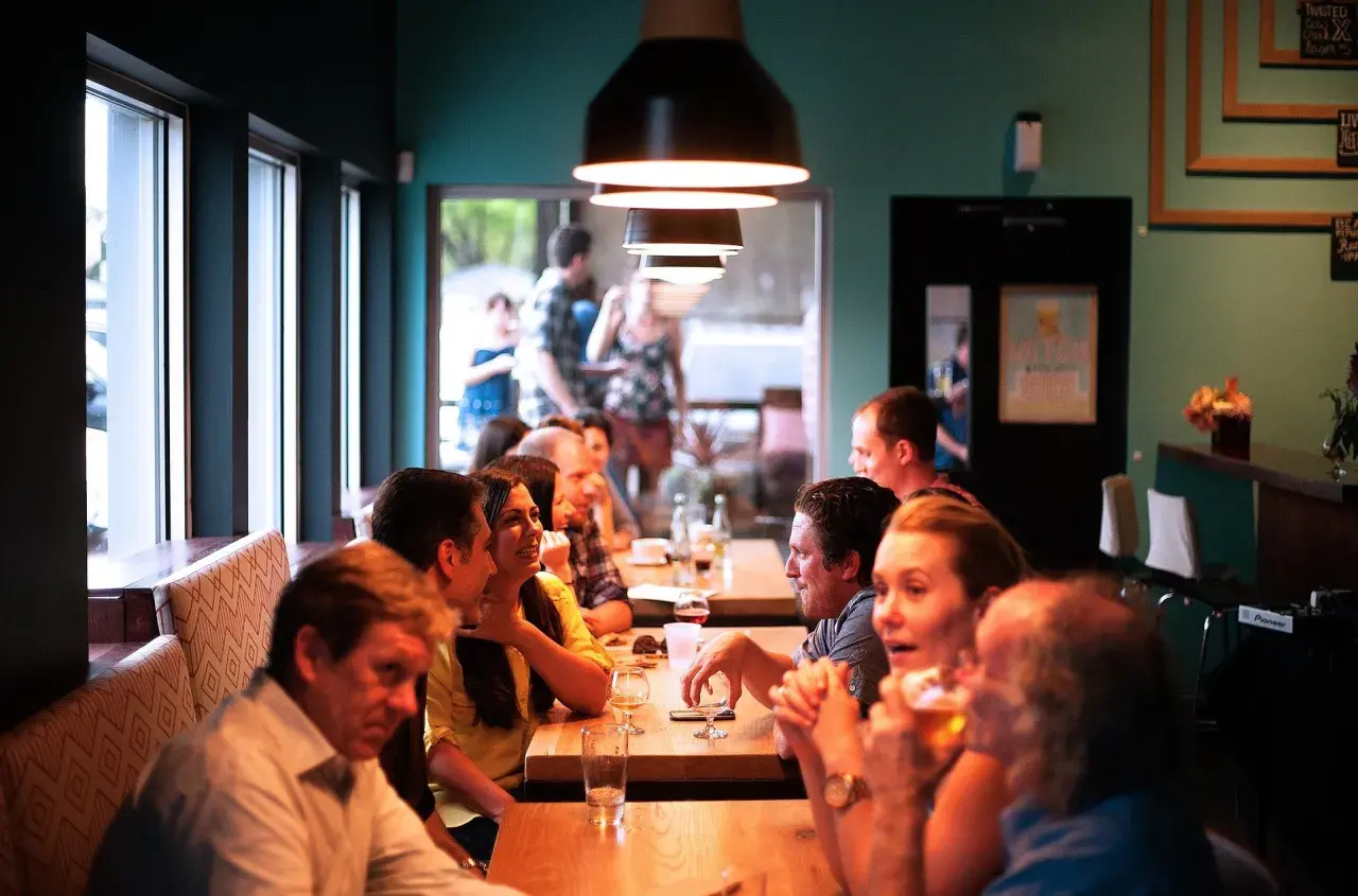 Ludzie siedzą przy stołach w przytulnym bistro, rozmawiają i piją.