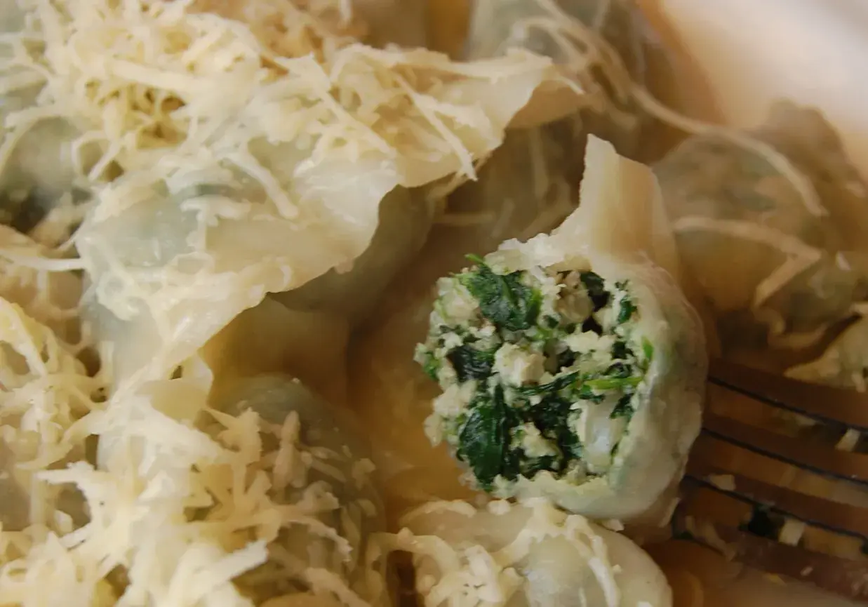 Pyszne pierogi z kurczakiem i szpinakiem, polane sosem i posypane startym serem. Jeden pieróg jest przekrojony, ukazując bogate nadzienie.
