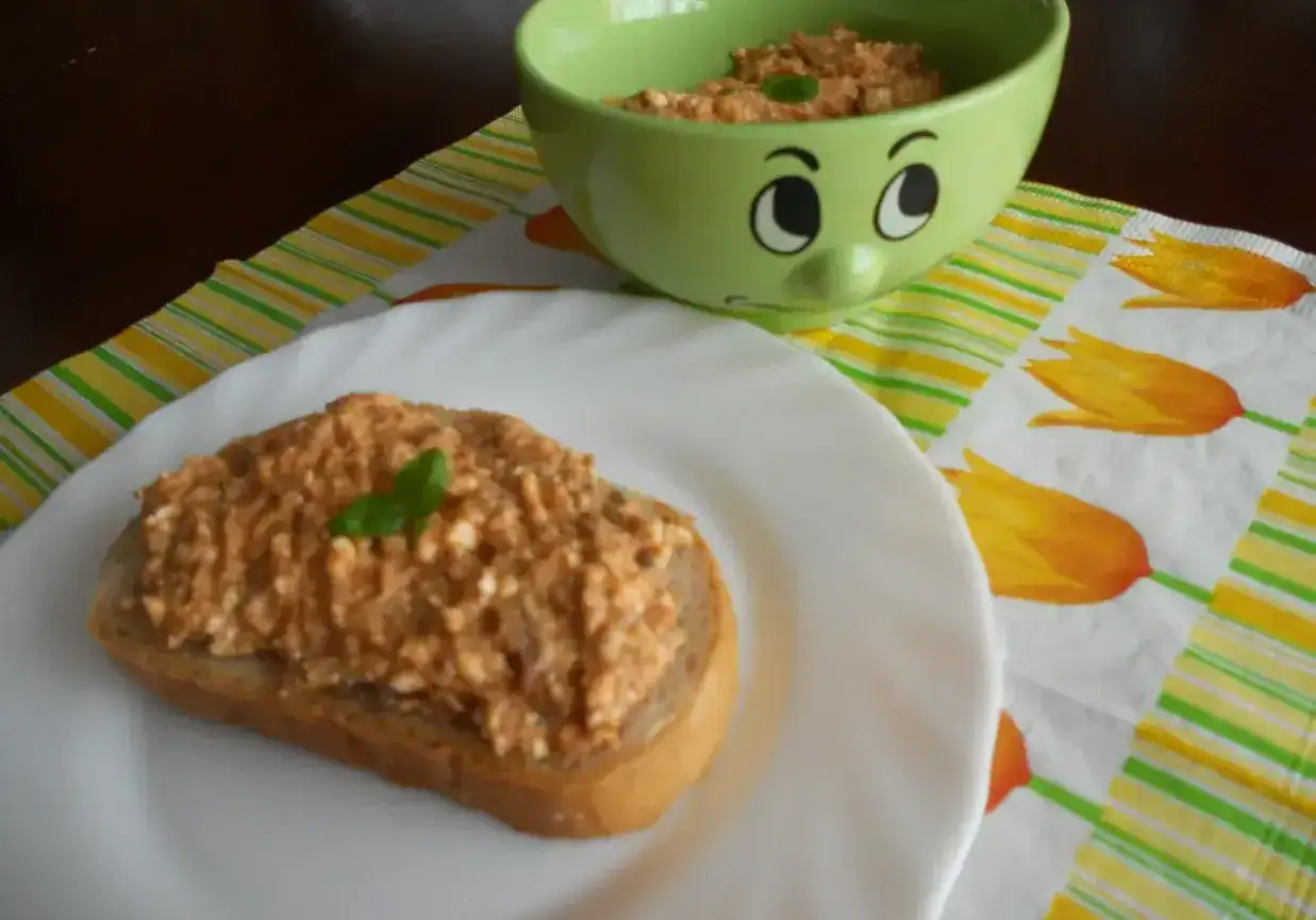 Pyszna pasta z makreli i twarogu na kromce chleba, ozdobiona listkiem bazylii. Obok miseczka z pastą i wesoła buźka.