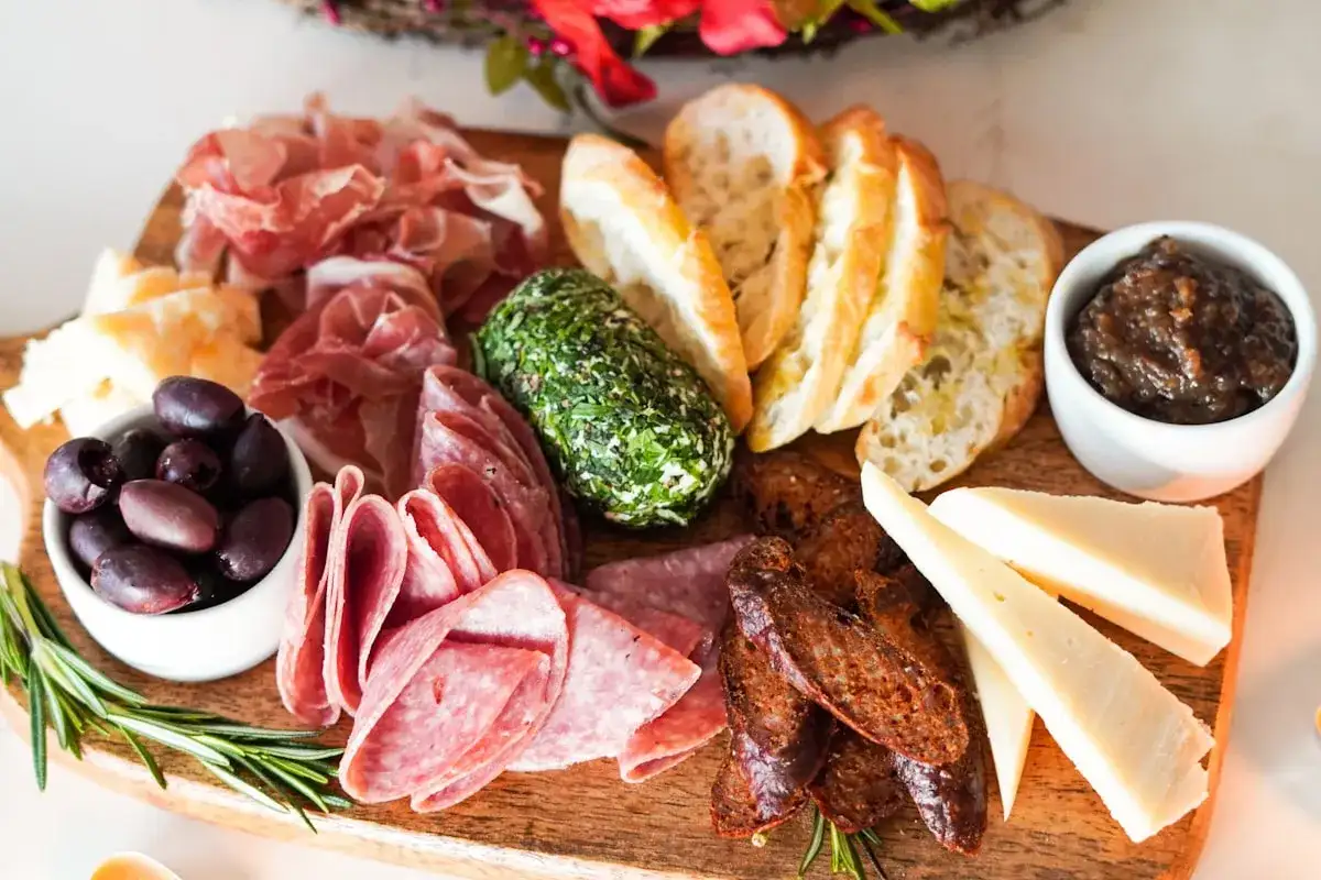 Apetyczna deska serów i wędlin: prosciutto, salami, oliwki, ser kozi w ziołach, bagietka, figowa konfitura i twarde sery.