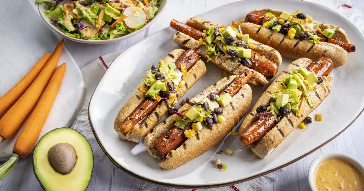 Pięć grillowanych hot dogów z marchewki, podanych w bułkach z awokado, czarną fasolą i kukurydzą. Obok miska sałatki i świeże marchewki.